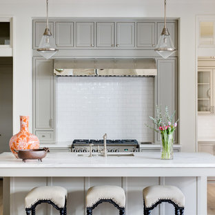 Cette photo montre une cuisine chic en U avec un évier encastré, un placard à porte shaker, des portes de placard grises, une crédence blanche, une crédence en carrelage métro, un électroménager en acier inoxydable, un sol en bois clair et un îlot central.