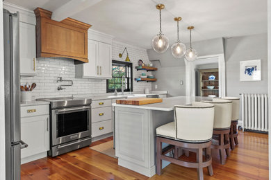 Example of a mid-sized classic l-shaped medium tone wood floor and beige floor enclosed kitchen design in Boston with a farmhouse sink, shaker cabinets, white cabinets, marble countertops, white backsplash, subway tile backsplash, stainless steel appliances, an island and white countertops