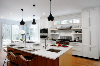 Example of a mid-sized trendy galley light wood floor and brown floor eat-in kitchen design in New York with a farmhouse sink, shaker cabinets, white cabinets, quartz countertops, white backsplash, subway tile backsplash, stainless steel appliances and an island