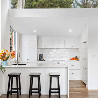Beach style l-shaped kitchen in Sydney with an undermount sink, shaker cabinets, white cabinets, white splashback, subway tile splashback, white appliances, medium hardwood floors, a peninsula, brown floor, white benchtop and vaulted.