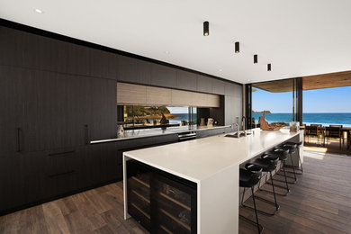 Пример оригинального дизайна: огромная параллельная кухня-гостиная в современном стиле с плоскими фасадами, темными деревянными фасадами, столешницей из акрилового камня, черным фартуком, зеркальным фартуком, черной техникой, островом и белой столешницей