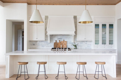 Example of a transitional galley light wood floor kitchen design in Salt Lake City with glass-front cabinets, white cabinets, white backsplash, an island, a farmhouse sink and stainless steel appliances