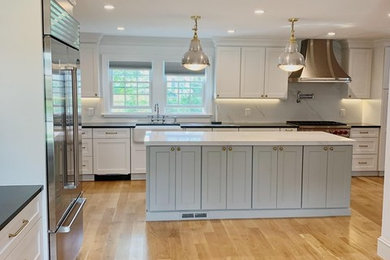 Inspiration for a large transitional l-shaped medium tone wood floor and beige floor eat-in kitchen remodel in New York with a farmhouse sink, recessed-panel cabinets, white cabinets, quartz countertops, white backsplash, stone slab backsplash, stainless steel appliances, an island and black countertops