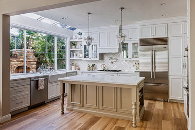 Example of a large classic light wood floor kitchen design with a farmhouse sink, recessed-panel cabinets, quartz countertops, white backsplash, porcelain backsplash, stainless steel appliances, an island and white countertops