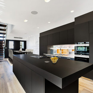 Photo of a contemporary galley eat-in kitchen in Sydney with an integrated sink, flat-panel cabinets, black cabinets, metallic splashback, black appliances, light hardwood floors, with island and black benchtop.