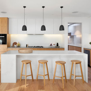 This is an example of a large contemporary l-shaped open plan kitchen in Melbourne with an undermount sink, flat-panel cabinets, medium wood cabinets, black appliances, medium hardwood floors, with island, brown floor, white benchtop, quartz benchtops and white splashback.