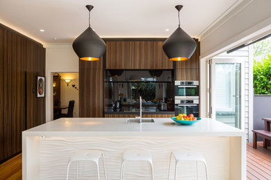 Moderne Küche in L-Form mit Unterbauwaschbecken, flächenbündigen Schrankfronten, dunklen Holzschränken, Küchenrückwand in Schwarz, Küchengeräten aus Edelstahl, braunem Holzboden, Kücheninsel, braunem Boden und weißer Arbeitsplatte in Auckland
