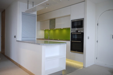 Medium sized modern galley open plan kitchen in London with an integrated sink, flat-panel cabinets, white cabinets, stainless steel worktops, green splashback, glass sheet splashback, stainless steel appliances, porcelain flooring and no island.