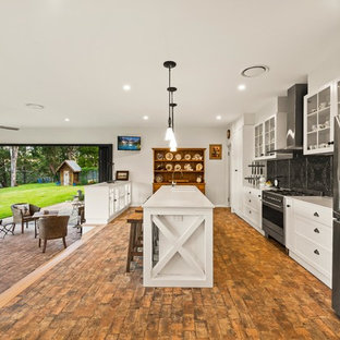 Photo of a country galley open plan kitchen in Other with shaker cabinets, white cabinets, black splashback, stainless steel appliances, brick floors, with island, brown floor and white benchtop.