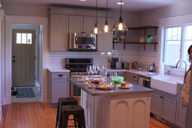 Photo of a medium sized farmhouse l-shaped kitchen/diner in Grand Rapids with a belfast sink, flat-panel cabinets, grey cabinets, engineered stone countertops, white splashback, ceramic splashback, stainless steel appliances, medium hardwood flooring and an island.
