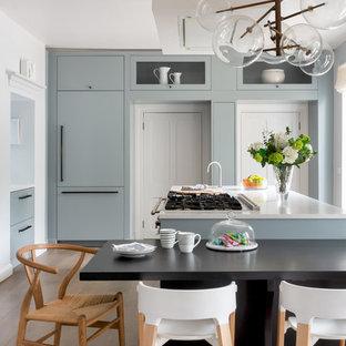 Traditional kitchen/diner in London with a double-bowl sink, flat-panel cabinets, blue cabinets, composite countertops, mirror splashback, stainless steel appliances, medium hardwood flooring, a breakfast bar, brown floors and white worktops.