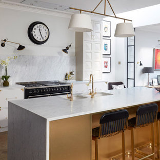 Photo of a large classic galley kitchen in London with a double-bowl sink, flat-panel cabinets, white cabinets, white splashback, stone slab splashback, black appliances, medium hardwood flooring, an island, brown floors and white worktops.
