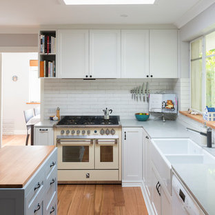 Inspiration for a transitional l-shaped separate kitchen in Canberra - Queanbeyan with a farmhouse sink, shaker cabinets, white cabinets, white splashback, brick splashback, white appliances, medium hardwood floors, with island, brown floor and grey benchtop.