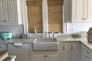Example of a mid-sized farmhouse dark wood floor and brown floor kitchen design in New York with a farmhouse sink, raised-panel cabinets, white cabinets, quartz countertops, white backsplash, an island and white countertops