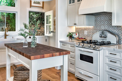 Example of a mid-sized beach style l-shaped medium tone wood floor and beige floor kitchen design in San Francisco with a farmhouse sink, white cabinets, quartz countertops, ceramic backsplash, white appliances, an island, white countertops, shaker cabinets and gray backsplash