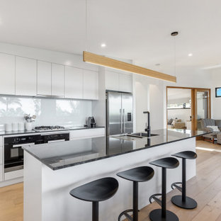Contemporary open plan kitchen in Sydney with a double-bowl sink, flat-panel cabinets, white cabinets, white splashback, glass sheet splashback, stainless steel appliances, light hardwood floors, with island and black benchtop.