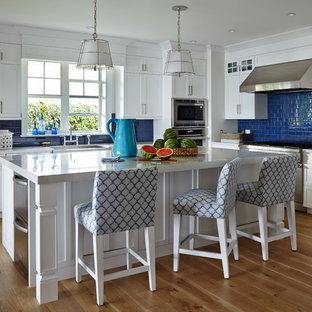Diseño de cocina marinera, de tamaño medio, con salpicadero azul, electrodomésticos de acero inoxidable, suelo de madera en tonos medios, puertas de armario blancas, armarios estilo shaker, salpicadero de azulejos tipo metro y una isla