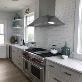 This is an example of a large country l-shaped kitchen/diner in Philadelphia with a belfast sink, beaded cabinets, grey cabinets, quartz worktops, white splashback, ceramic splashback, stainless steel appliances, medium hardwood flooring, an island and orange floors.