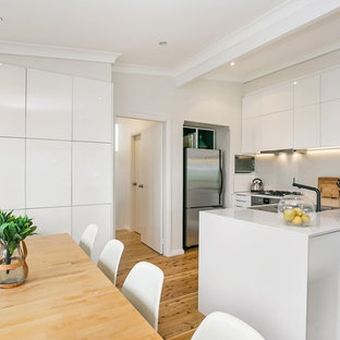 Beach style u-shaped kitchen in Central Coast with a double-bowl sink, flat-panel cabinets, white cabinets, white splashback, stainless steel appliances, medium hardwood floors, a peninsula, brown floor and white benchtop.