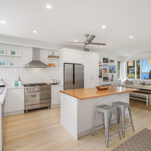 Photo of a beach style l-shaped kitchen in Central Coast with a drop-in sink, flat-panel cabinets, white cabinets, white splashback, subway tile splashback, stainless steel appliances, light hardwood floors, with island, beige floor and white benchtop.