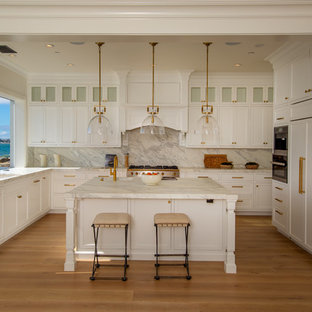 Beach style u-shaped kitchen in Los Angeles with an integrated sink, recessed-panel cabinets, white cabinets, white splashback, window splashback, panelled appliances, medium hardwood floors, with island, brown floor and white benchtop.