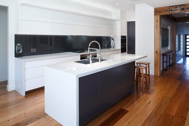Moderne Wohnküche in L-Form mit weißen Schränken, Quarzit-Arbeitsplatte, Küchenrückwand in Braun, Kücheninsel, Unterbauwaschbecken, Glasrückwand, braunem Holzboden, braunem Boden, flächenbündigen Schrankfronten, schwarzen Elektrogeräten und weißer Arbeitsplatte in Sydney