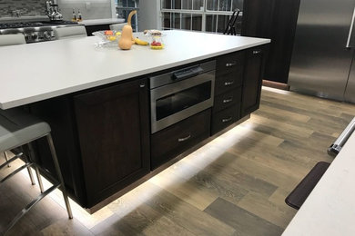Mid-sized trendy medium tone wood floor and brown floor eat-in kitchen photo in Dallas with shaker cabinets, dark wood cabinets, quartzite countertops, gray backsplash, stainless steel appliances, an island, white countertops, an undermount sink and glass tile backsplash