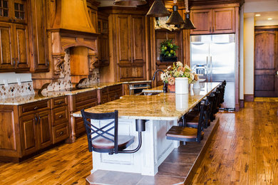 Example of a large classic galley medium tone wood floor eat-in kitchen design in Salt Lake City with a farmhouse sink, raised-panel cabinets, medium tone wood cabinets, granite countertops, brown backsplash, ceramic backsplash, stainless steel appliances and an island