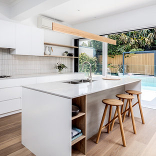 Beach style galley kitchen in Sydney with an undermount sink, flat-panel cabinets, white cabinets, white splashback, mosaic tile splashback, medium hardwood floors, with island, brown floor and white benchtop.