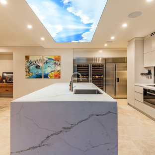 This is an example of a contemporary open plan kitchen in Sydney with a single-bowl sink, flat-panel cabinets, beige cabinets, metallic splashback, mirror splashback, black appliances, with island, beige floor and beige benchtop.