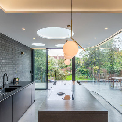 Mid-sized minimalist galley concrete floor and gray floor eat-in kitchen photo in London with an integrated sink, flat-panel cabinets, black cabinets, solid surface countertops, brick backsplash, black appliances and an island