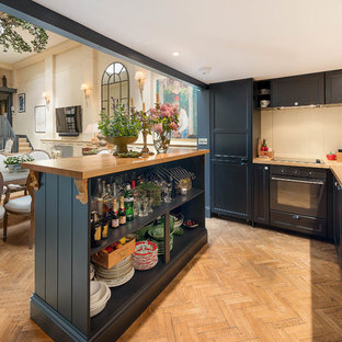 Medium sized traditional l-shaped kitchen in London with shaker cabinets, blue cabinets, wood worktops, beige splashback, black appliances, light hardwood flooring, an island, brown floors and brown worktops.