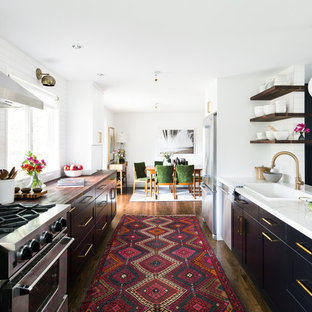 Foto de cocina comedor de galera, clásica renovada, con fregadero encastrado, armarios con paneles lisos, puertas de armario negras, salpicadero blanco, electrodomésticos de acero inoxidable, suelo de madera en tonos medios y península