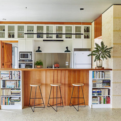 Open concept kitchen - mid-sized transitional galley concrete floor and gray floor open concept kitchen idea in Other with glass-front cabinets, white cabinets, wood countertops, metallic backsplash, stainless steel appliances, metal backsplash, a peninsula and brown countertops