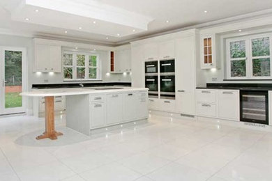 This is an example of an expansive classic l-shaped kitchen in Surrey with a submerged sink, flat-panel cabinets, white cabinets, marble worktops, black splashback, marble splashback, black appliances, porcelain flooring, an island and white floors.