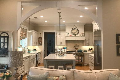 Example of a large classic u-shaped medium tone wood floor and brown floor open concept kitchen design in Miami with an island, a farmhouse sink, shaker cabinets, white cabinets, quartz countertops, gray backsplash, subway tile backsplash, stainless steel appliances and white countertops