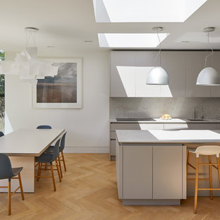 Photo of a scandi galley kitchen/diner in London with a submerged sink, flat-panel cabinets, grey cabinets, grey splashback, black appliances, light hardwood flooring, an island, beige floors and grey worktops.