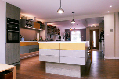 Medium sized contemporary galley open plan kitchen in London with an integrated sink, flat-panel cabinets, yellow cabinets, laminate countertops, metallic splashback, stainless steel appliances, medium hardwood flooring, an island, brown floors and white worktops.