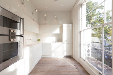 Moderne Küche ohne Insel in L-Form mit Doppelwaschbecken, flächenbündigen Schrankfronten, weißen Schränken, Küchengeräten aus Edelstahl und hellem Holzboden in London