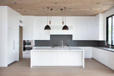 Large contemporary l-shaped open plan kitchen in Los Angeles with a submerged sink, flat-panel cabinets, white cabinets, limestone worktops, grey splashback, stone slab splashback, stainless steel appliances, light hardwood flooring, an island and grey worktops.