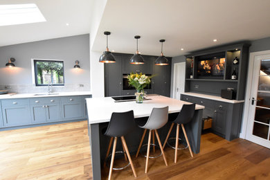 Photo of a large modern l-shaped open plan kitchen in Wiltshire with an integrated sink, shaker cabinets, grey cabinets, engineered stone countertops, grey splashback, stone tiled splashback, black appliances, medium hardwood flooring, an island and white worktops.