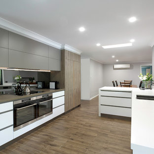 This is an example of a large contemporary galley eat-in kitchen in Brisbane with flat-panel cabinets, medium hardwood floors, brown floor, a double-bowl sink, grey cabinets, black splashback, glass sheet splashback, black appliances, a peninsula and white benchtop.