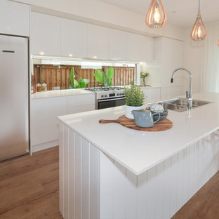 This is an example of a large beach style galley kitchen in Sunshine Coast with a drop-in sink, white cabinets, quartz benchtops, stainless steel appliances, with island, brown floor, white benchtop, flat-panel cabinets, window splashback and medium hardwood floors.