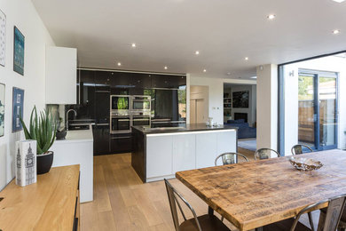 This is an example of a medium sized modern l-shaped open plan kitchen with an integrated sink, flat-panel cabinets, black cabinets, laminate countertops, integrated appliances, light hardwood flooring, an island and black worktops.