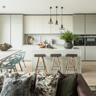 Design ideas for a contemporary open plan kitchen in London with flat-panel cabinets, white cabinets, white splashback, black appliances, light hardwood flooring, an island and white worktops.