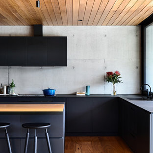 Contemporary l-shaped kitchen in Melbourne with a drop-in sink, flat-panel cabinets, black cabinets, grey splashback, cement tile splashback, black appliances, with island, black benchtop, medium hardwood floors and brown floor.