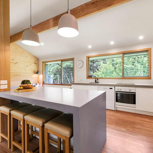 Photo of a beach style kitchen in Melbourne with a single-bowl sink, flat-panel cabinets, white cabinets, white appliances, medium hardwood floors, with island, grey benchtop and exposed beam.