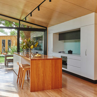Photo of a contemporary galley kitchen in Melbourne with a double-bowl sink, flat-panel cabinets, white cabinets, wood benchtops, blue splashback, glass sheet splashback, panelled appliances, medium hardwood floors, with island and brown floor.