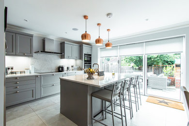 This is an example of a large classic l-shaped kitchen/diner in Surrey with an integrated sink, shaker cabinets, grey cabinets, quartz worktops, white splashback, stone slab splashback, black appliances, white worktops, an island and grey floors.