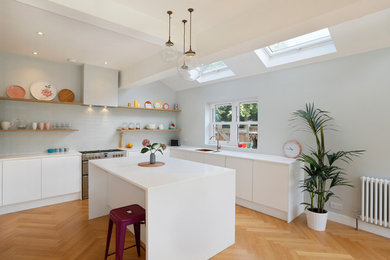 Photo of a contemporary l-shaped kitchen in Other with a submerged sink, flat-panel cabinets, white cabinets, grey splashback, metro tiled splashback, stainless steel appliances, medium hardwood flooring, an island, brown floors and white worktops.